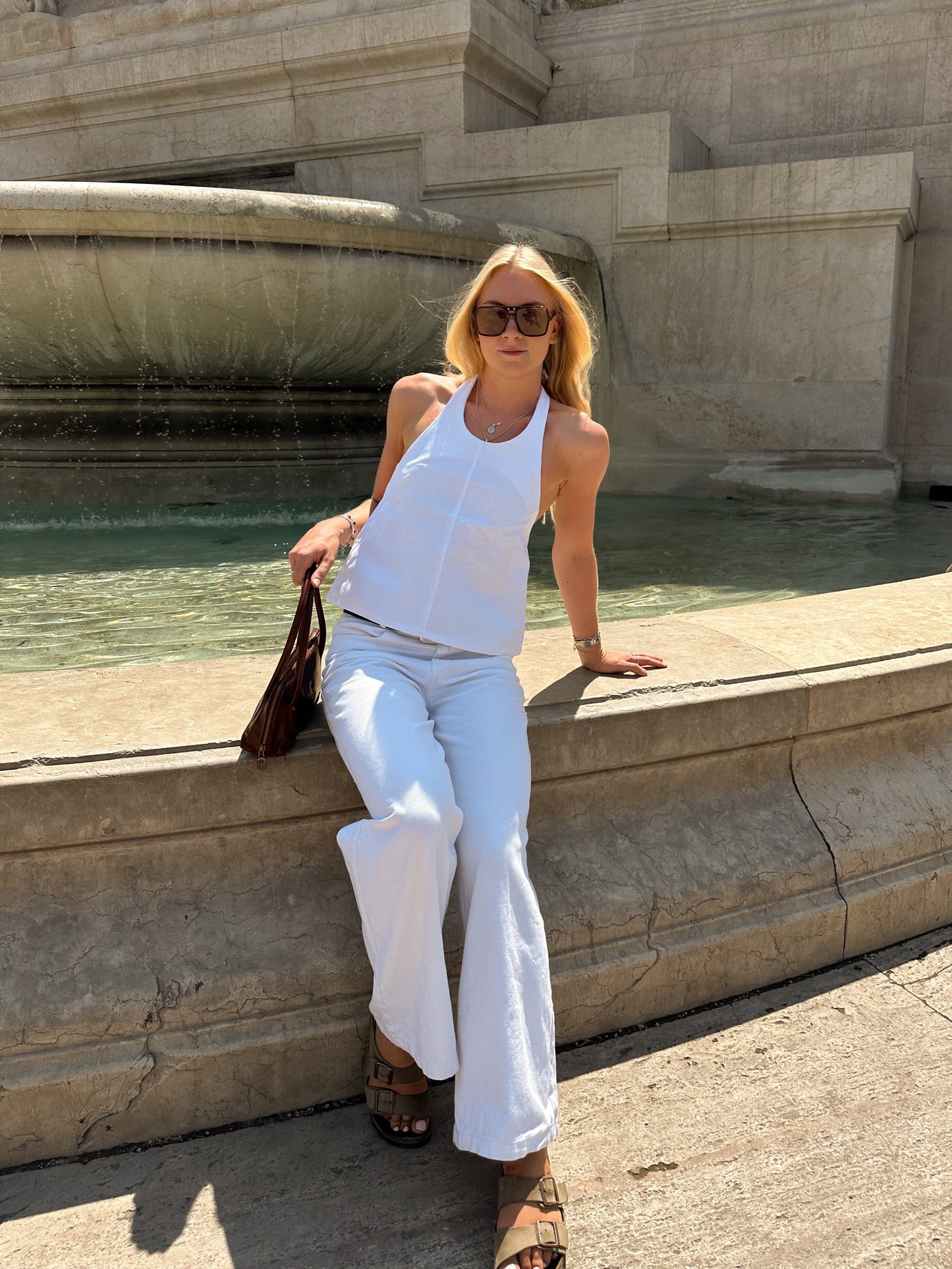 Woman in white halter neck top and white wide leg jeans sitting by a fountain with classical architecture