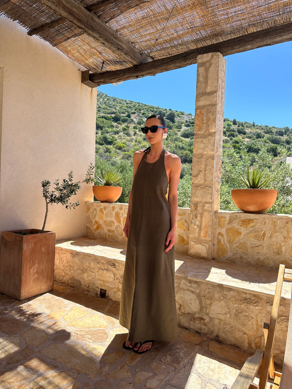 Woman in a long dress standing in a scenic outdoor setting with stone walls and greenery.
