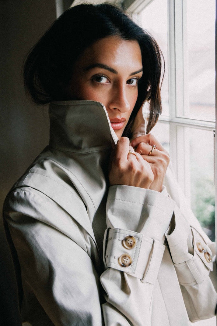 A woman wearing a light beige trench coat stands by a window, pulling the collar up with both hands. The coat has structured shoulders, flap pockets with decorative buttons, and a classic tailored silhouette.