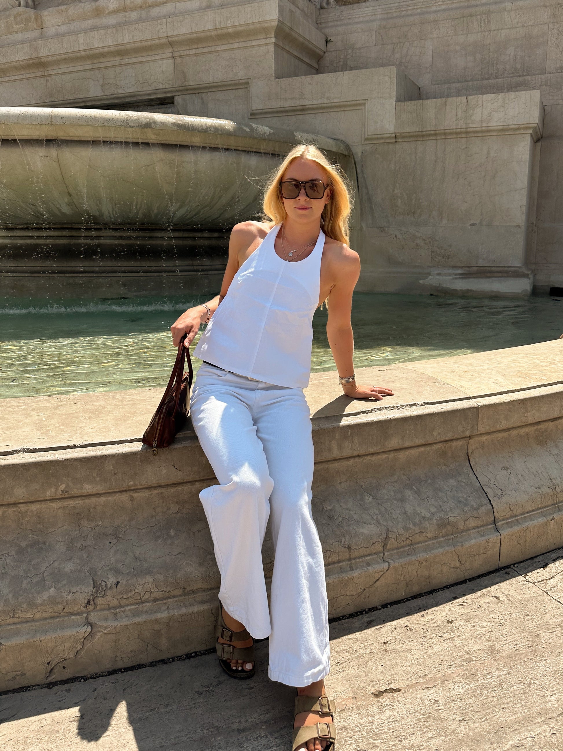 Woman in white halter neck top and white wide leg jeans sitting by a fountain with classical architecture